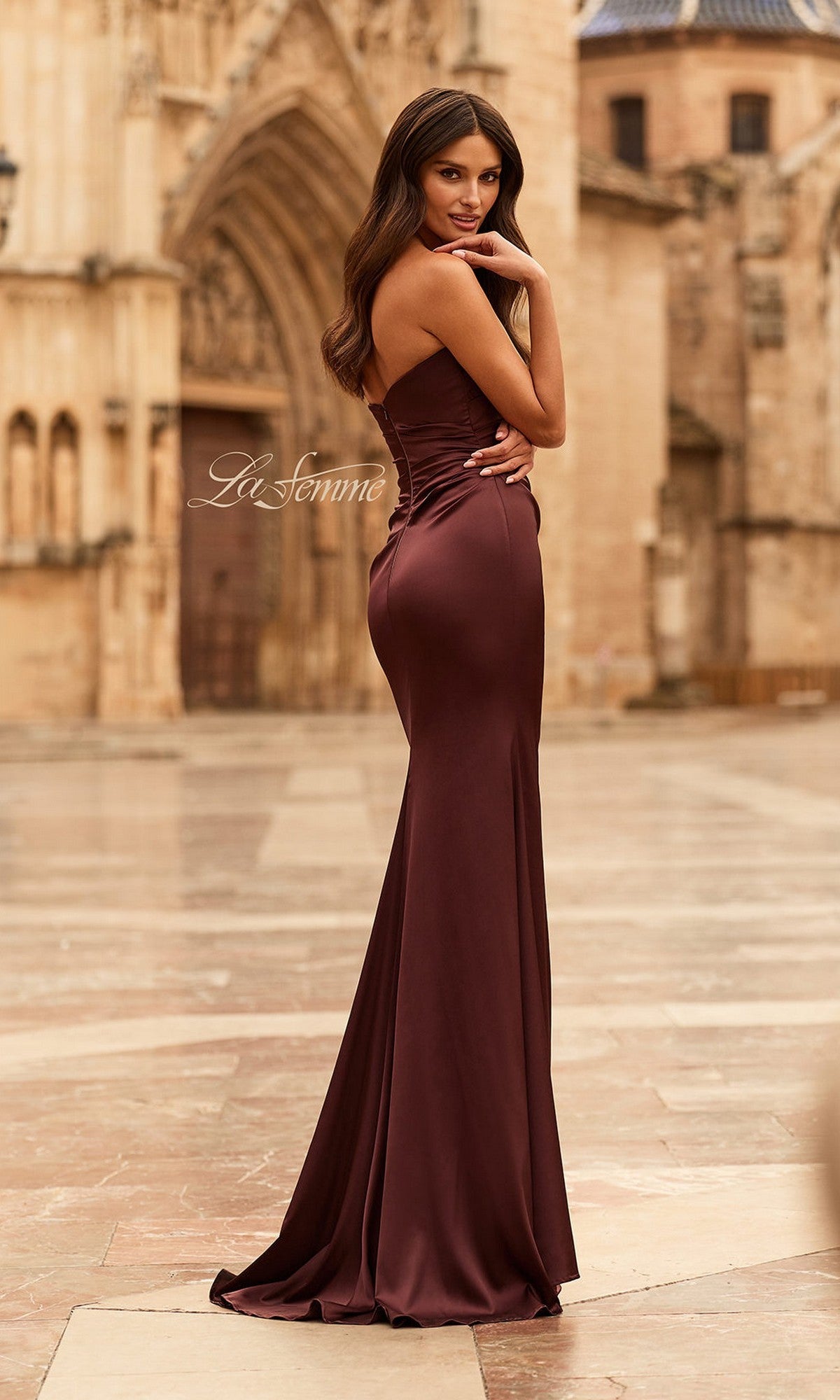 A woman wears the La Femme 33670 long prom dress with a ruched bodice, standing outdoors on stone pavement with historic architecture in the background.